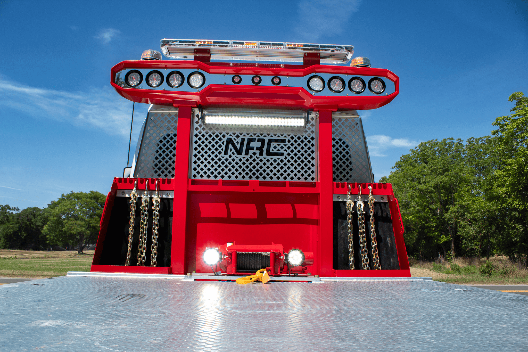 Back view of a red tow truck equipped with LED light bars and chains, set against a blue sky and green trees.