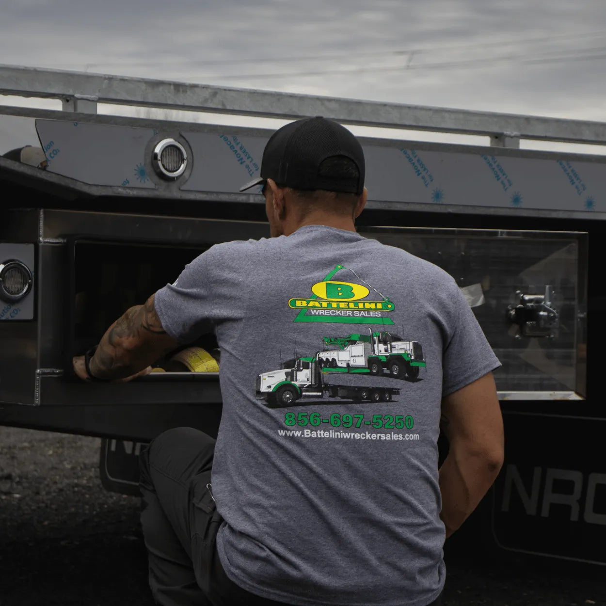 Person wearing a gray t-shirt with a truck dealership logo, kneeling next to a large vehicle.