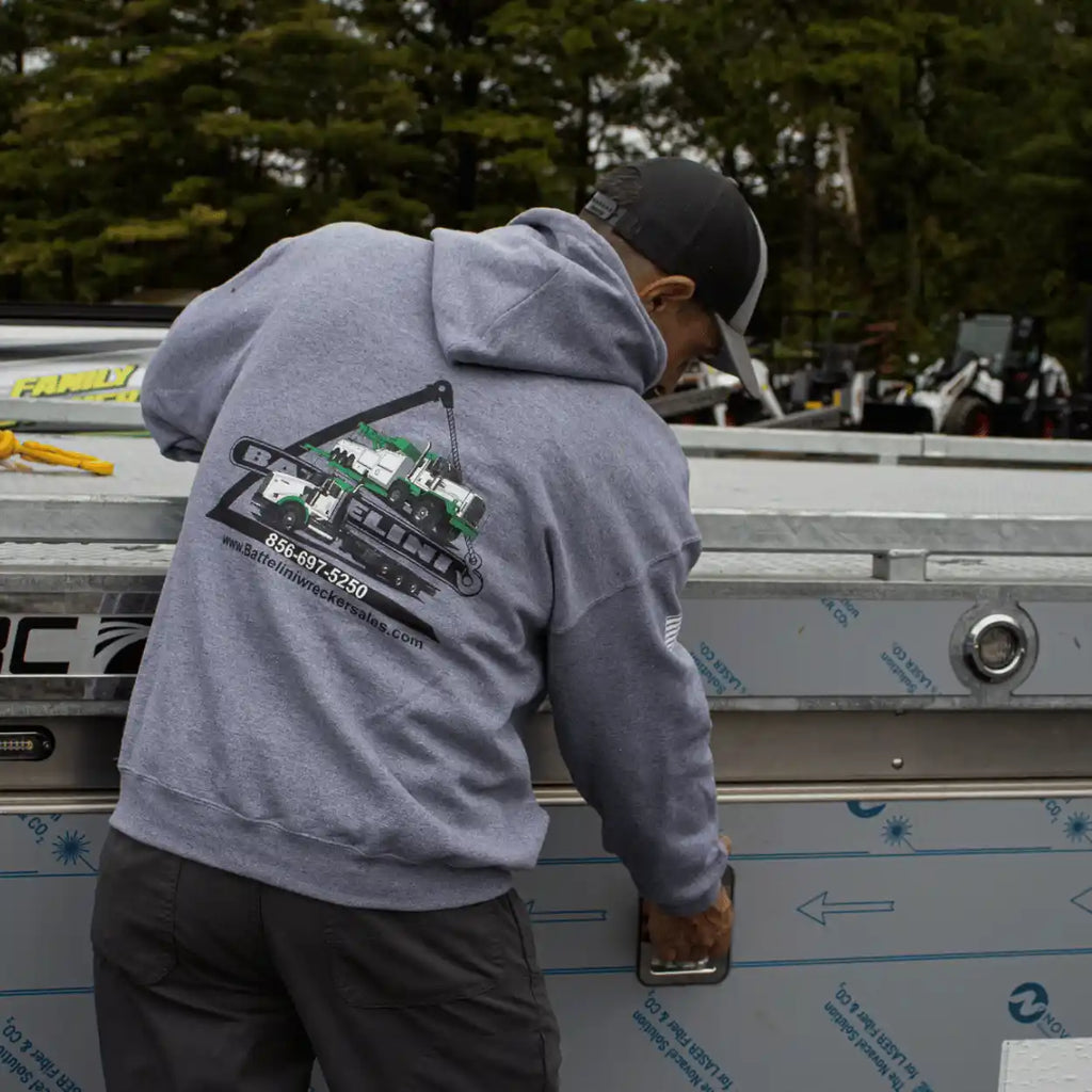 Person wearing a gray battelini trucking hoodie with a logo, working on a metal surface with trees in the background.