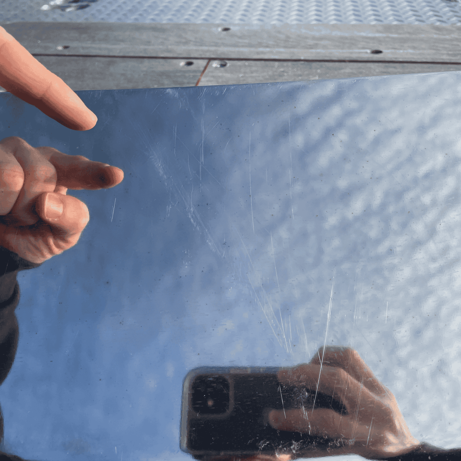 Close-up of minor surface scratches on a used stainless steel Paccar sun visor for Kenworth W900. Reflection of hand and phone visible on the surface.